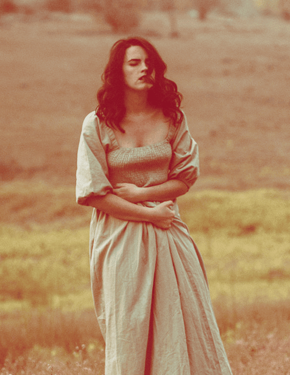 A woman standing in a sunlit field with her arms crossed, gazing inward